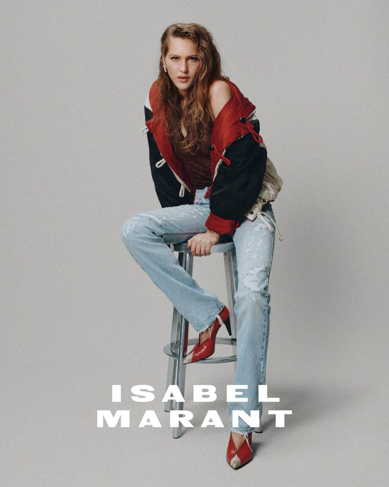 A stylish, fair-skinned woman with long, wavy red hair poses in studio, wearing a red-and-black jacket, distressed jeans and red heels.