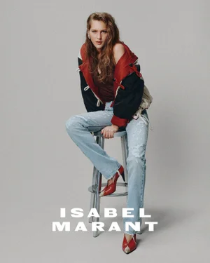 Model seated on a metal stool wearing a red and black jacket, distressed denim jeans, and red heels against a grey seamless background