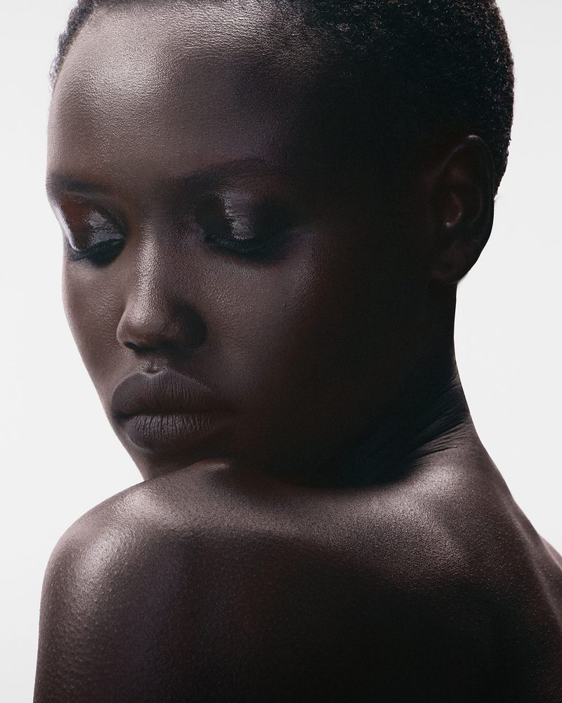 Close-up portrait of a dark-skinned adult female with short hair, gazing down with a serious expression in a brightly lit studio setting.
