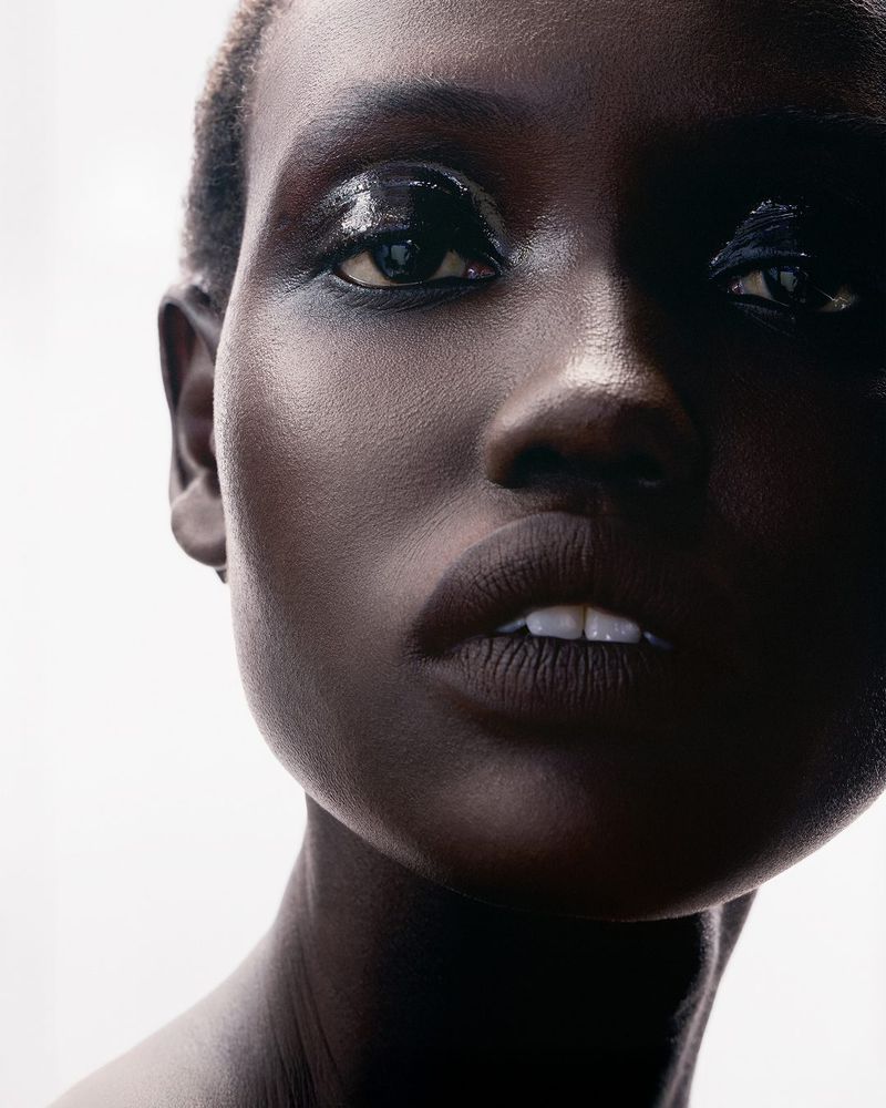 Close-up studio shot of a Black woman with dark hair and glossy makeup, showcasing beauty and skincare in a minimalist setting against a white background.