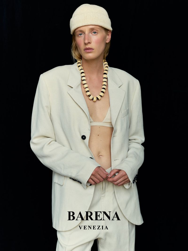 Fashion shot of an adult female wearing an off-white suit, matching beanie, and beaded necklace against a black backdrop in a studio.
