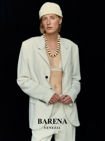 Fashion shot of an adult female wearing an off-white suit, matching beanie, and beaded necklace against a black backdrop in a studio.