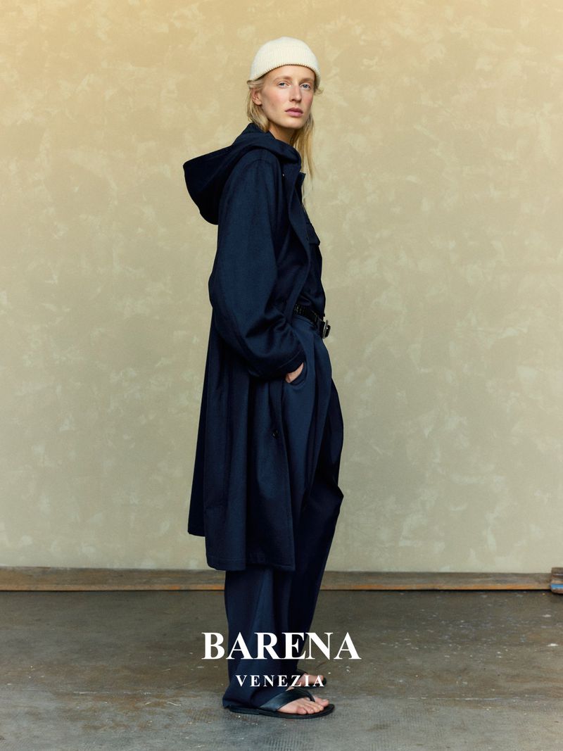 An adult female poses in a studio wearing a navy hooded coat, navy pants, a cream cap, and sandals. Neutral stucco background. 