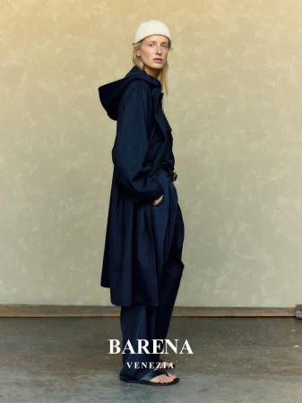 An adult female poses in a studio wearing a navy hooded coat, navy pants, a cream cap, and sandals. Neutral stucco background. 