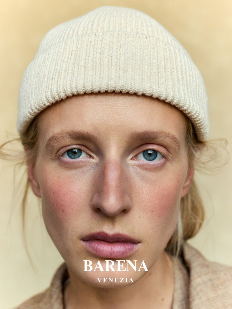 Close-up portrait of a young fair-skinned woman wearing a cream-colored knit beanie and a tan jacket, against a cream backdrop.