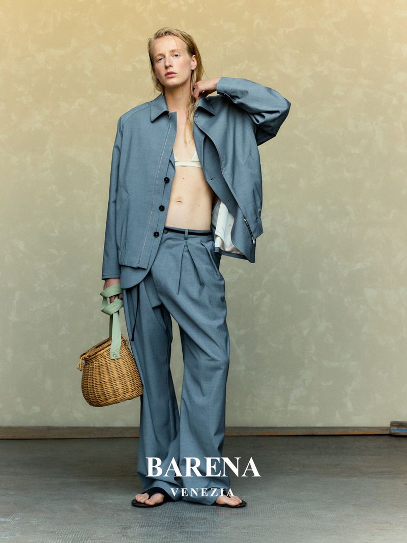 Fashion editorial shot: A slim adult woman wears a light blue oversized suit, bralette top, and flip-flops. She's holding a woven basket.