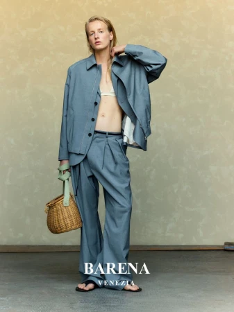 Model wearing a blue jacket and matching trousers holding a woven basket bag against a textured backdrop