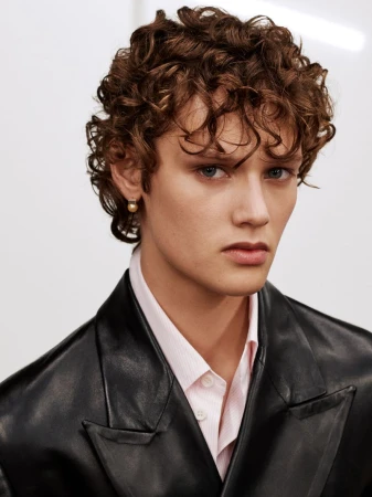 Portrait of a man with curly brown hair wearing a black leather blazer and pink collared shirt against a white wall.
