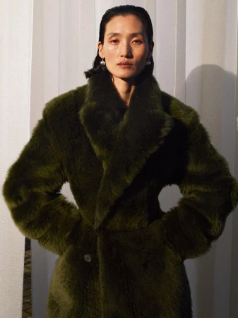 A woman with sleek dark hair wears a thick, textured olive green faux fur coat, looking directly at the camera in a portrait shot.