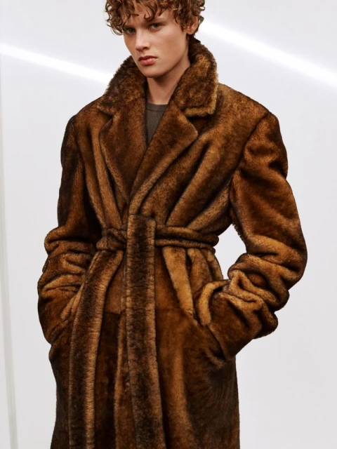 Male model poses wearing a brown faux fur trench coat with a belt in a studio setting with soft, diffused lighting and a neutral background.
