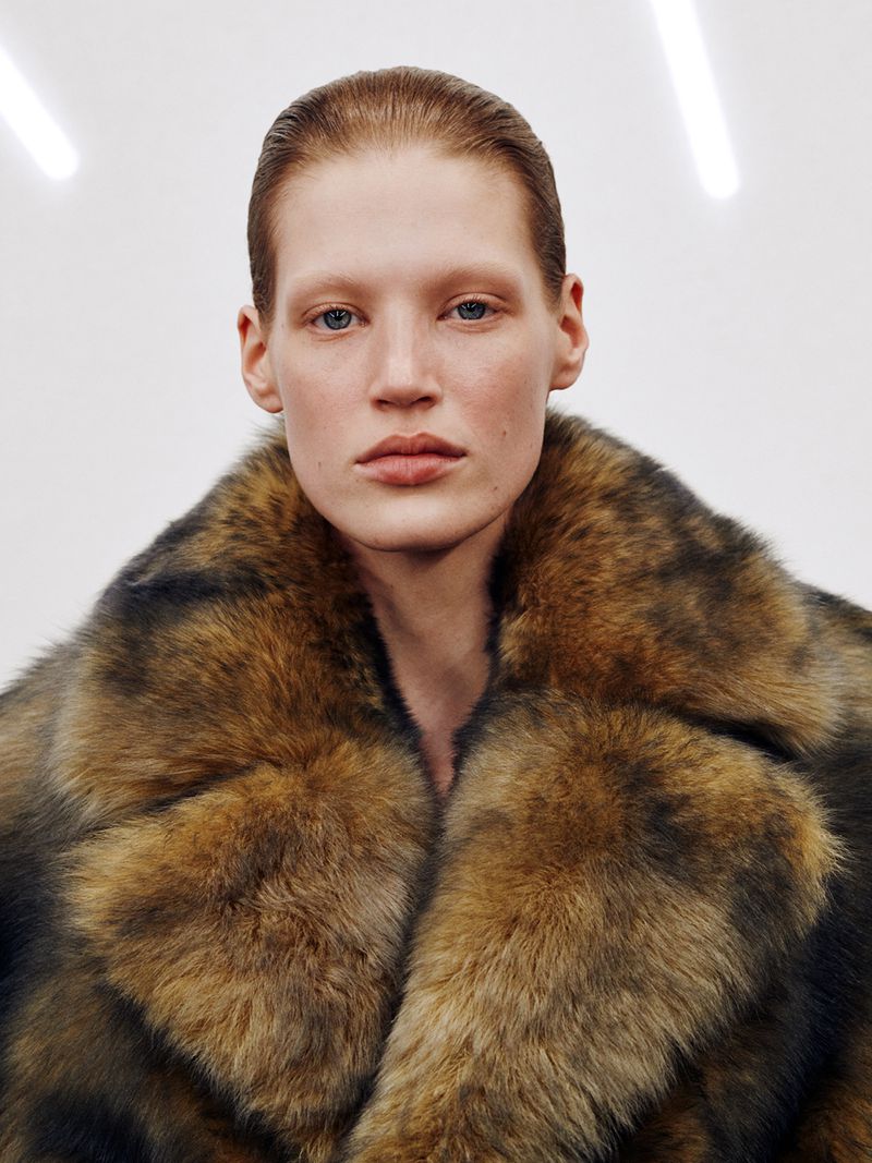 Close-up portrait of a light-skinned adult woman wearing a brown fur coat against a white background with bright fluorescent lighting.