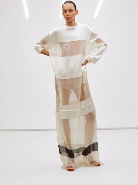A slender woman poses in a studio wearing a long, layered, sheer dress. She is fair-skinned and wears her hair pulled back.