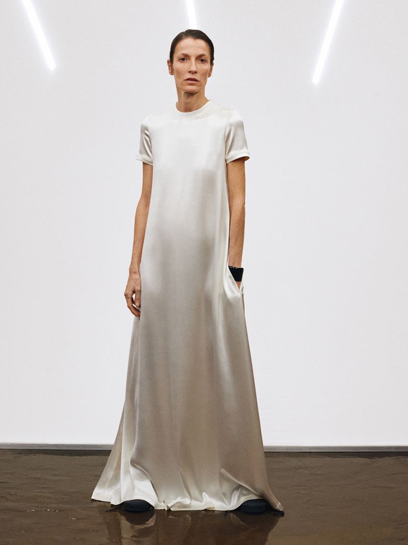 Elegant woman in long cream dress with short sleeves and pockets stands against a bright white background with angled neon lights.