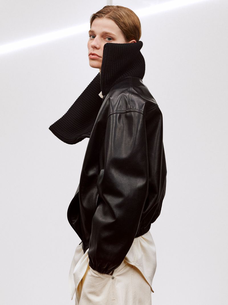 Fashionable person wearing black leather jacket and cream pants against a white backdrop, striking a pose.