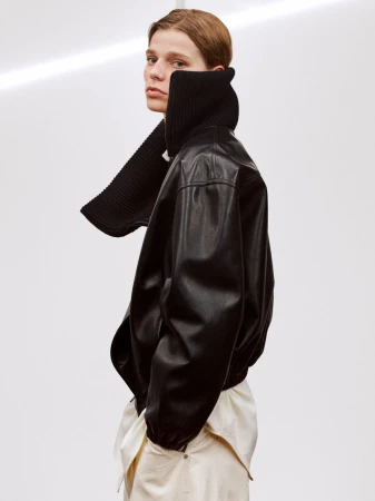 Fashionable person wearing black leather jacket and cream pants against a white backdrop, striking a pose.