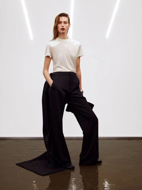 Fashion editorial shot of a slender, fair-skinned woman with wet hair, wearing a T-shirt and long, wide-leg black trousers in studio.