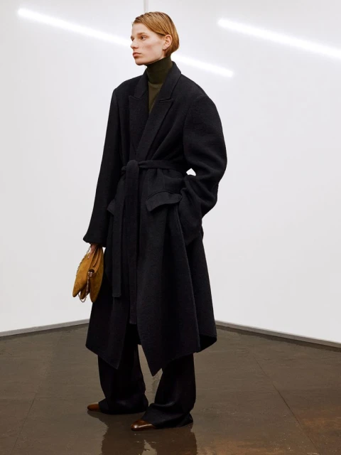 A fashionable woman in a long, dark coat poses in a minimalist studio. She's holding a suede purse, complemented by the warm, neutral tones.