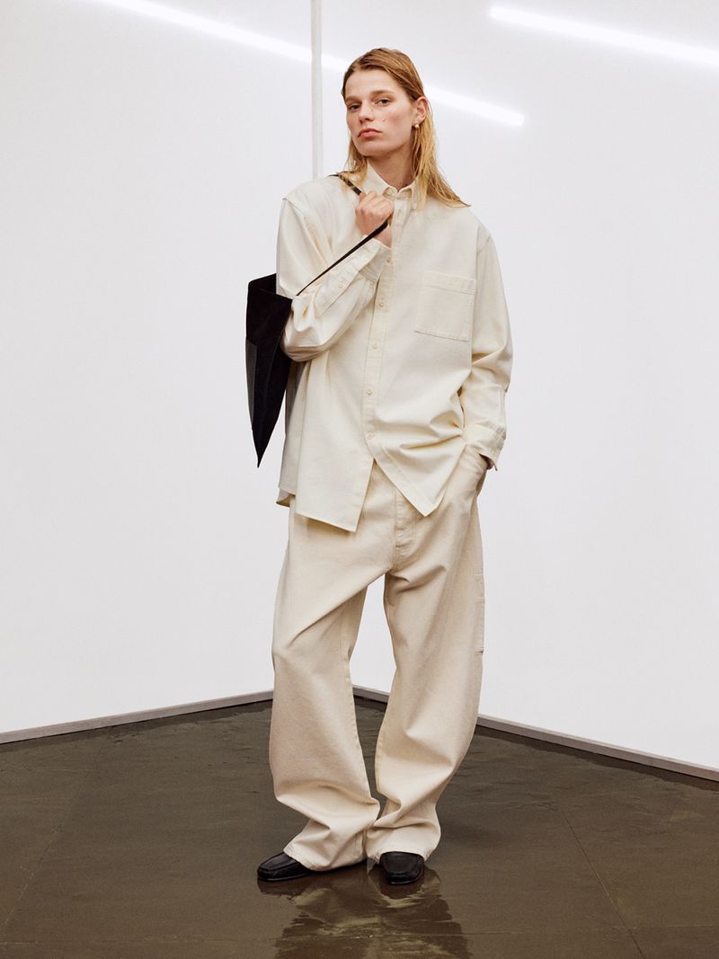 An adult fashion model wears an oversized button-down shirt, wide-leg pants, and flat shoes in a minimalist white corner photo studio.