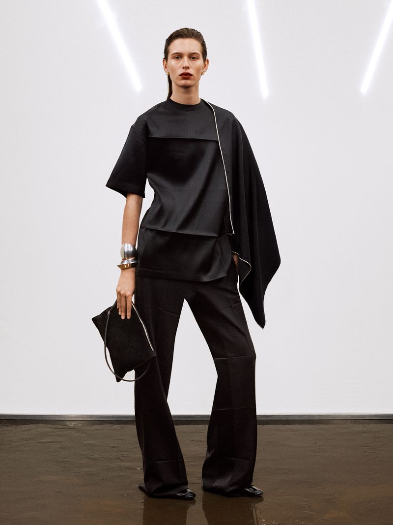 Fashion model wearing a black two-piece outfit in front of a white wall with studio lighting. She is holding a matching black clutch.
