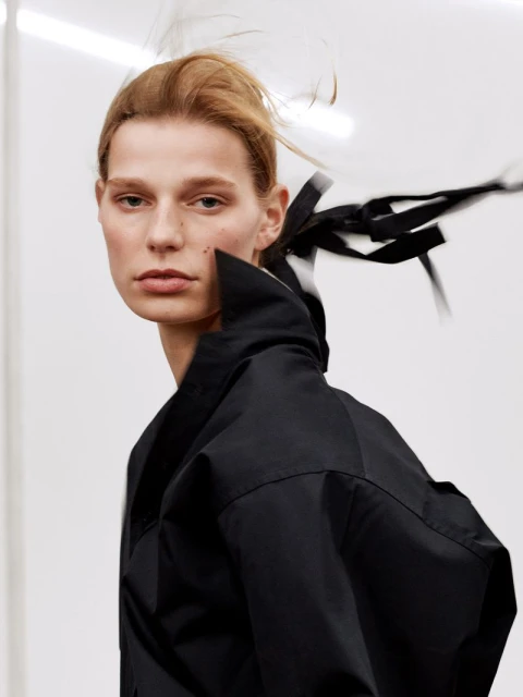 Close-up fashion portrait of an adult female model with fair skin and reddish blonde hair wearing a black coat in a bright studio.