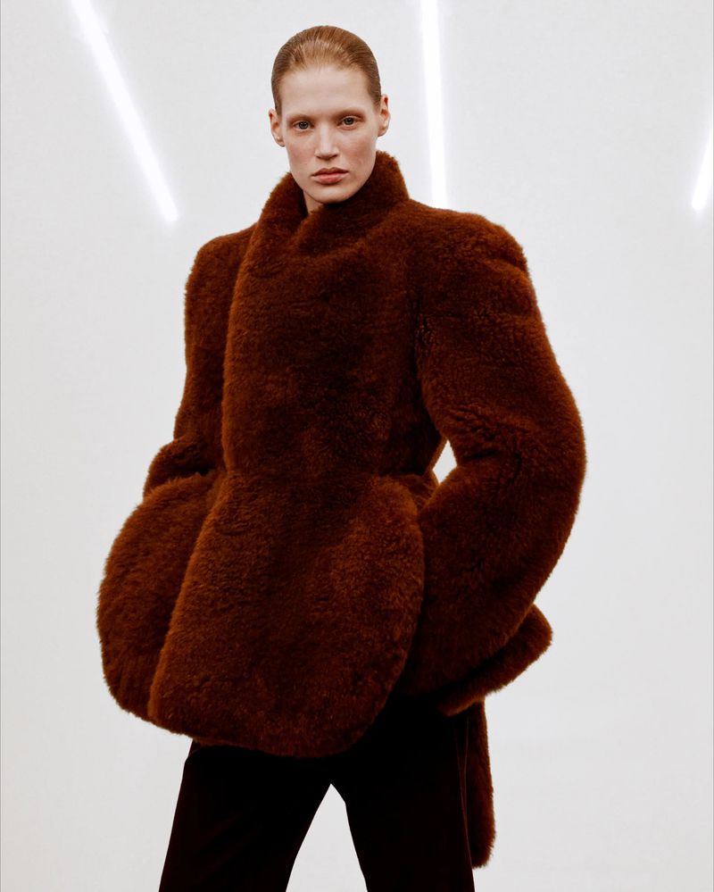 Model wearing a structured brown textured coat against a bright white seamless background with vertical light streaks.