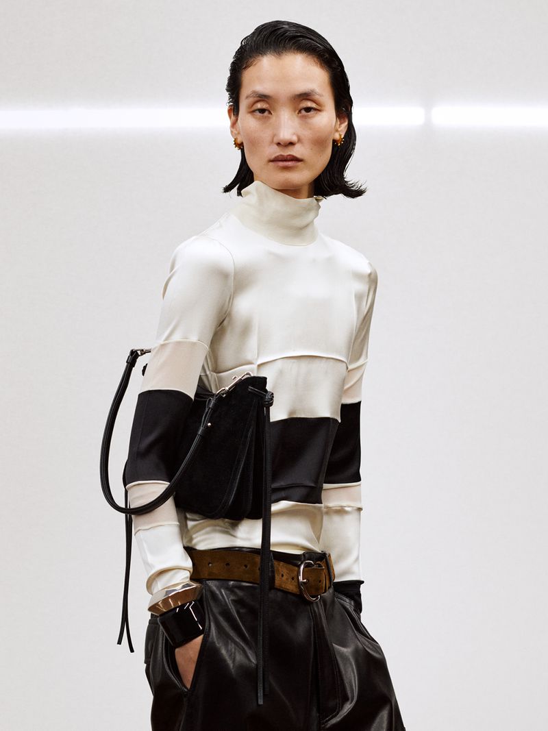 Fashion portrait of a woman with black handbag wearing a white and black top and leather pants in a bright studio setting.