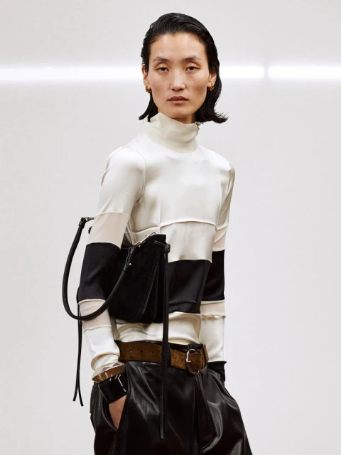 Fashion portrait of a woman with black handbag wearing a white and black top and leather pants in a bright studio setting.