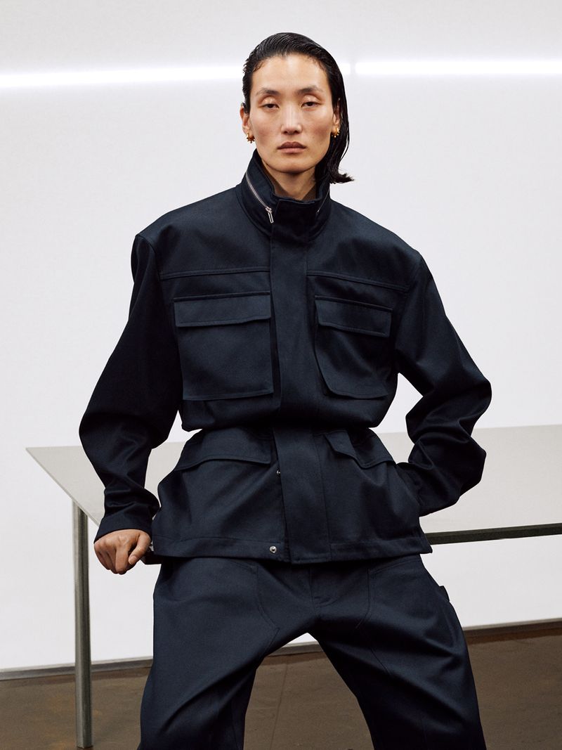 Model wearing a dark utility jacket and matching pants posed against a plain white studio wall and table