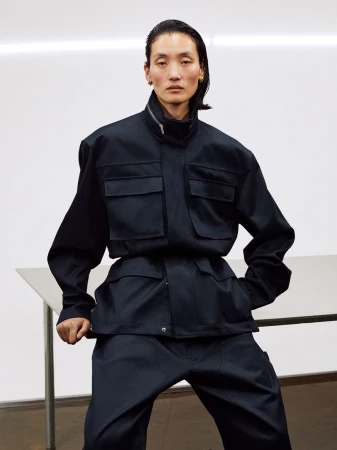 Model wearing a dark utility jacket and matching pants posed against a plain white studio wall and table