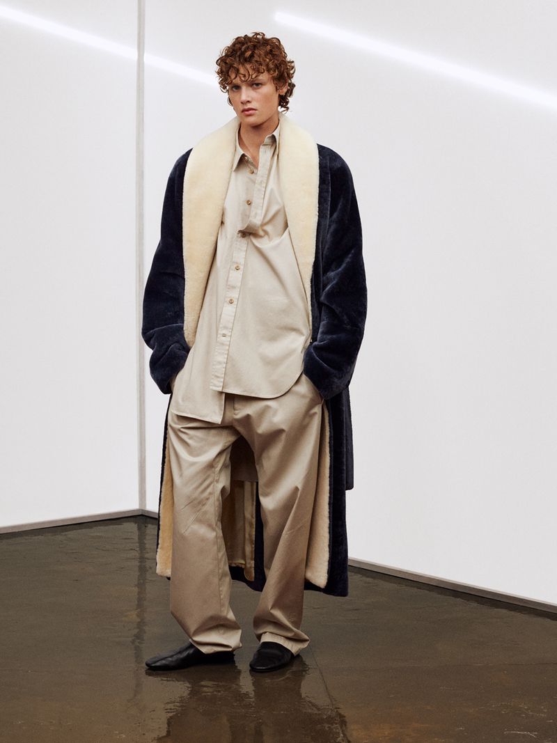 Fashionable adult man wearing oversized beige shirt and pants with a large navy blue faux fur coat with a cream colored lining.