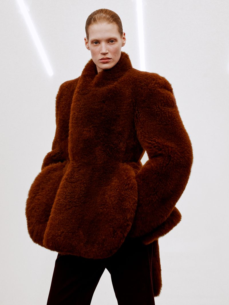 A fair-skinned model poses indoors wearing a brown fur coat and black pants against a bright white background with horizontal lighting.