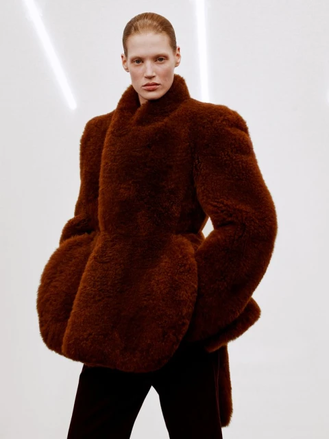 A fair-skinned model poses indoors wearing a brown fur coat and black pants against a bright white background with horizontal lighting.