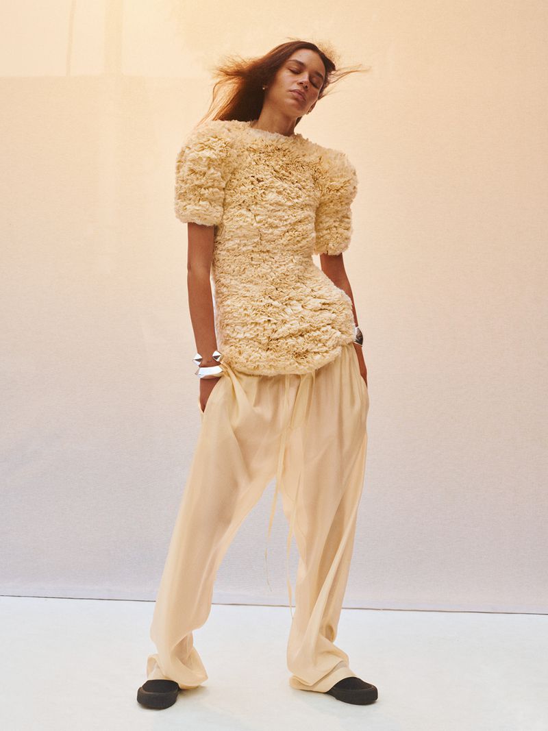 Fashion photo of a female model in textured top and light pants, posing against a soft gradient backdrop in a studio setting.