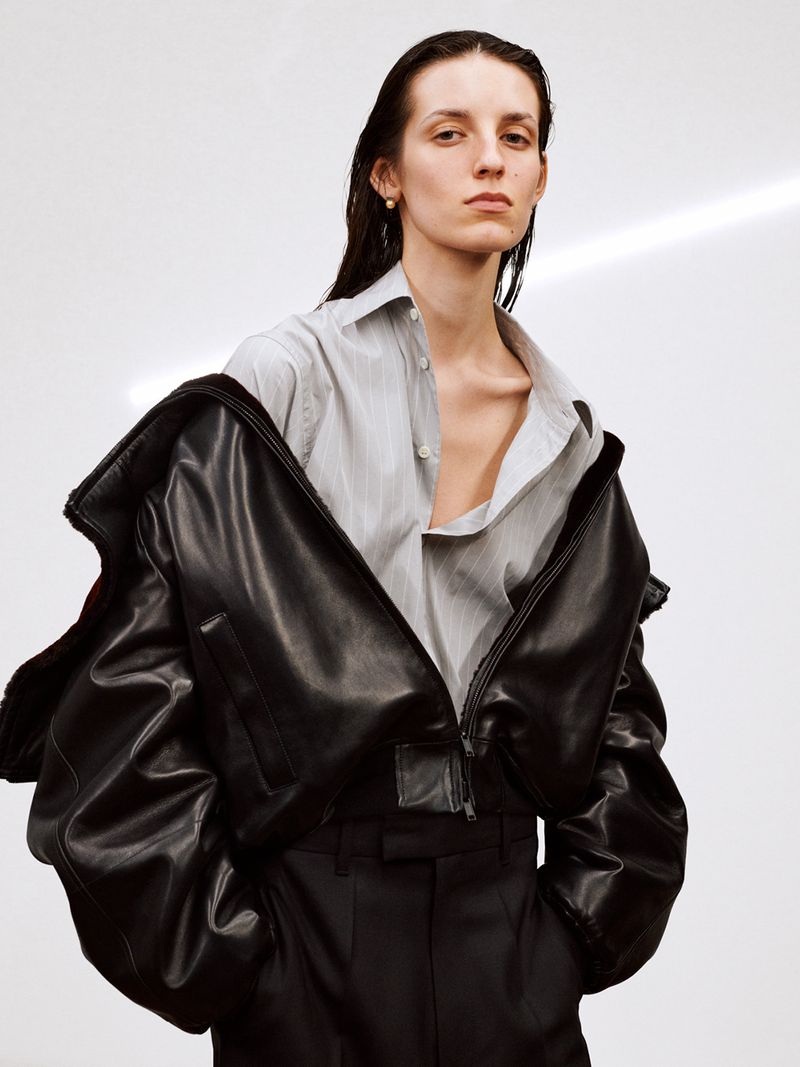 Model wearing a black leather jacket and striped shirt against a plain white studio backdrop.