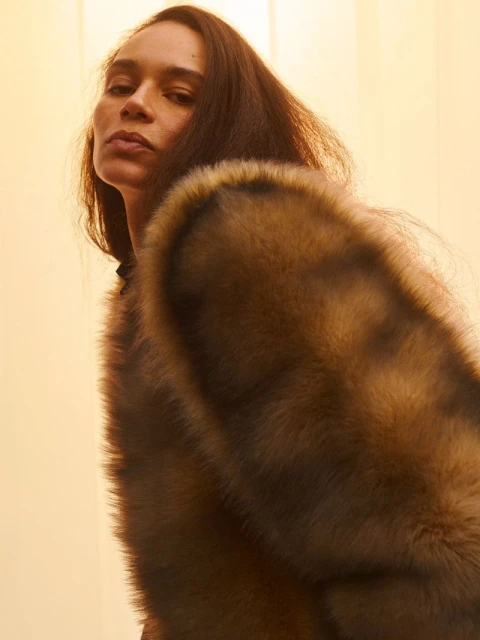 A woman with long brown hair looks up at the camera while wearing a brown fur coat against a pale yellow gradient background.