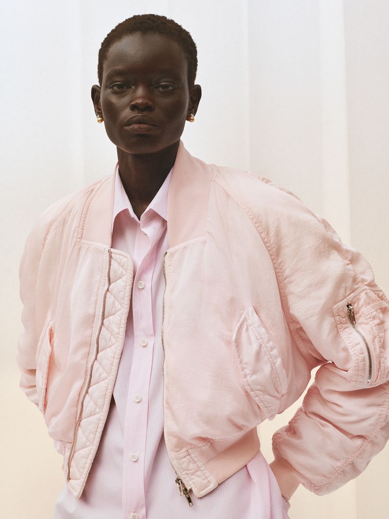 Close-up shot of a dark-skinned female model in a light pink jacket and shirt, posing against a neutral background in a fashion editorial.