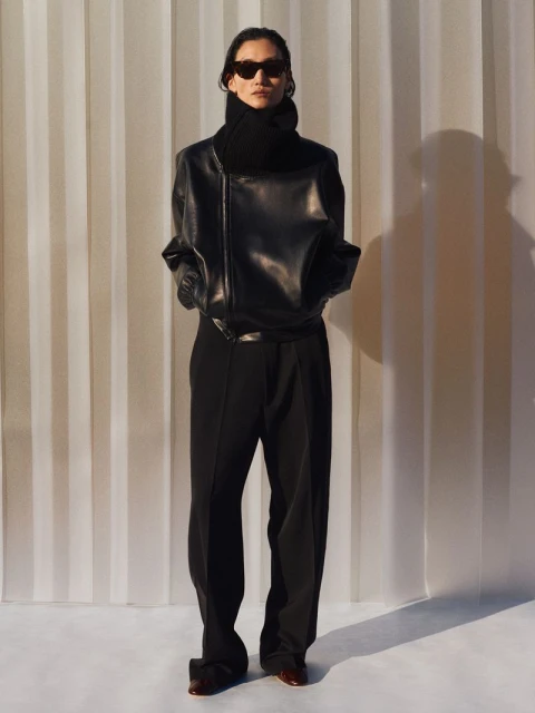 A person in a black leather jacket, black pants, and sunglasses stands in front of a light-colored curtain backdrop in a fashion studio shot.