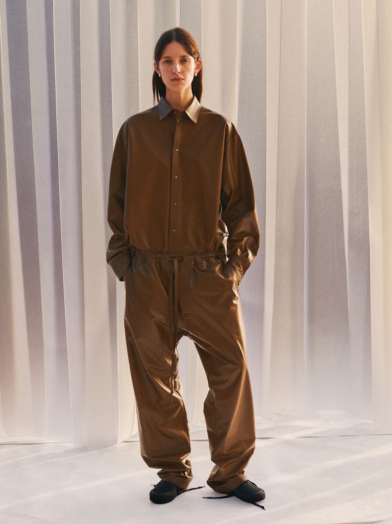 Woman in brown jumpsuit poses in front of white curtain backdrop, fashionable studio portrait, full body shot, fashion editorial style.