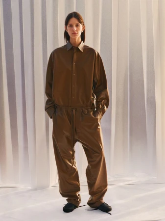 Model wearing a brown button-up shirt and matching trousers against a pale vertical curtain backdrop