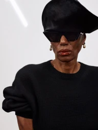 Model wearing dark sunglasses, black hat, and black knit sweater against a plain white background
