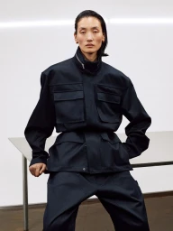 Model wearing a dark utility jacket and matching pants posed against a plain white studio wall and table