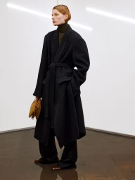 Model wearing a black belted coat and brown handbag against a clean white backdrop with overhead lighting.