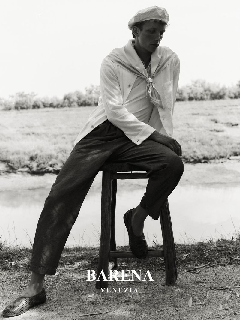Monochrome portrait of a young man wearing a sailor's hat and shirt, sitting on a stool in a rural environment, showcasing a classic fashion style.