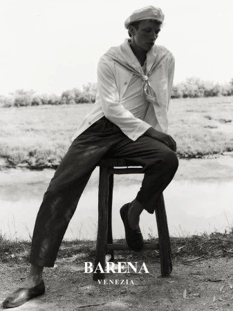 Monochrome portrait of a young man wearing a sailor's hat and shirt, sitting on a stool in a rural environment, showcasing a classic fashion style.