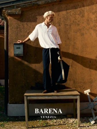 A fair-skinned male model poses outdoors, wearing a white shirt and hat, dark pants, standing on a wooden table with a muted wall background.