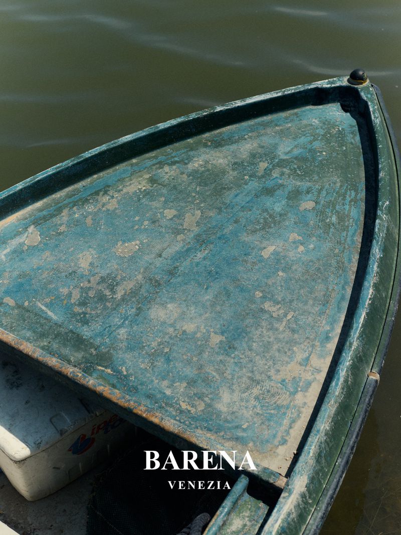 Close-up of a blue-green rowboat on calm water, featuring weathered paint and the brand BARENA VENEZIA.