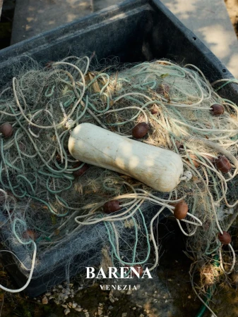Close-up of a fishing net in a grey container with white and brown floats. BARENA VENEZIA logo visible in the lower left.