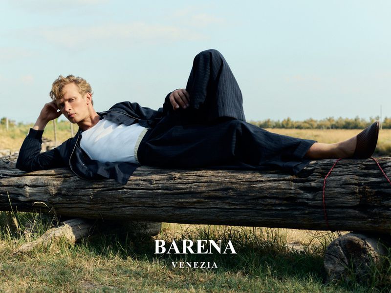 A blond male fashion model poses on a log outdoors wearing a white t-shirt, dark blue pants, jacket, and brown shoes against a blue sky.