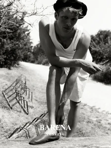 Model wearing a white tank top and dark cap leaning outdoors with stacked metal frames in the background for Barena Venezia.