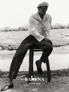 Model seated on a wooden stool wearing a white jacket and cap outdoors by water for Barena Venezia.